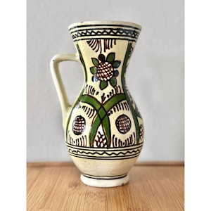 May include: A hand-painted ceramic pitcher with a cream-colored base and intricate floral and geometric designs in green, brown, and black. The pitcher has a handle and a narrow neck, showcasing a traditional folk art style.