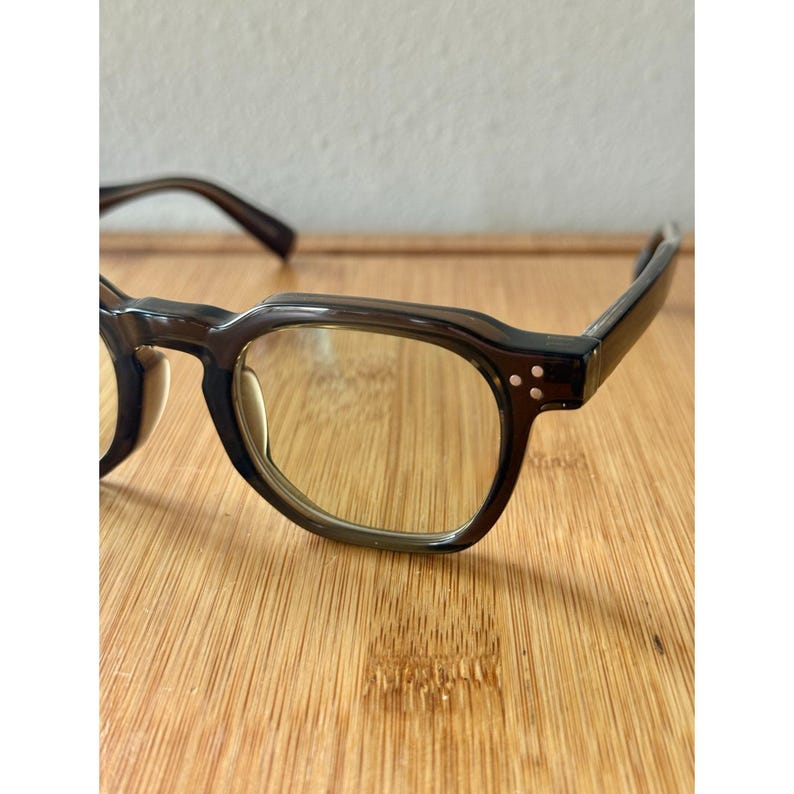 May include: A pair of dark brown eyeglasses with a square frame. The glasses have clear lenses and small, round rivets on the temples. The frames are a translucent brown color.