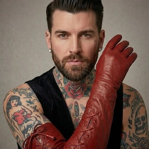 May include: A pair of long, deep red leather gloves featuring a lace-up design that extends from the wrist up the forearm. The gloves include thin matching red laces threaded through metal eyelets, creating a crisscross pattern along the length.
