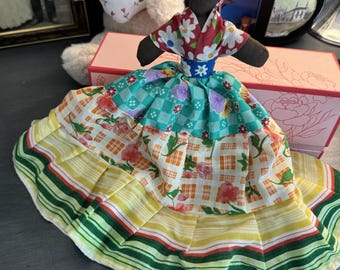 May include: A handmade fabric doll with a dark brown complexion, wearing a multi-layered dress featuring a red and white floral bodice, a blue waist band, and a skirt with green checkered, orange plaid, and yellow striped fabric panels.