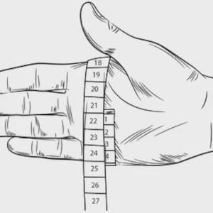 May include: A black and white line drawing of a hand with a measuring tape wrapped around the palm. The tape shows numerical markings ranging from 18 to 27, indicating a method for measuring hand circumference for sizing purposes in a clear illustration.