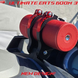 May include: A red portable Bluetooth speaker mounted on a black metal bracket. The bracket is attached to a white surface. The speaker has a black strap and a black and red design. The text "ULTIMATE EATS BOOM 3" is visible at the top of the image. The text "NEW DESIGN!" is visible at the bottom of the image.