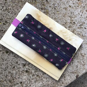 Op de afbeelding: Een zwarte etui met een bloemenpatroon en een roze elastiek, voorzien van een rits.