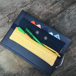 Op de afbeelding: Een marineblauw notitieboek met een geel patroon etui en een marineblauwe pennenhouder. De etui heeft een gele rits en bevat een groene pen en een markeerstift. De pennenhouder heeft witte cirkelontwerpen.