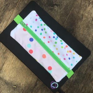 Könnte beinhalten: Ein rechteckiges Federmäppchen mit einem grünen Reißverschluss und einer grünen Stofflasche. Das Etui besteht aus zwei Stoffen: einem mit rosa Hintergrund und bunten Punkten und einem mit weißem Hintergrund und bunten Punkten. Das Etui liegt auf einem schwarzen Notizbuch.