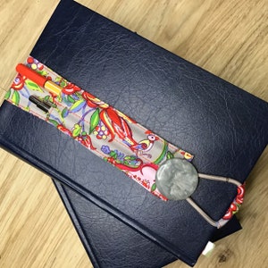Op de afbeelding: Een blauw notitieboek met een bloemenstof bladwijzer. De bladwijzer heeft een zilveren penhouder en een grijze knop.