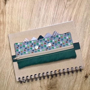 Könnte beinhalten: Ein handgefertigtes Federmäppchen mit grünem Boden und einem gemusterten Oberteil mit Dreiecken in verschiedenen Farben. Das Etui hat einen Reißverschluss und zwei weiße kreisförmige Augen. Es ist an einem Spiralnotizbuch mit einem Gummiband befestigt.