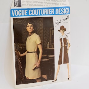 May include: A vintage Vogue Couturier Design sewing pattern featuring a dress design by Sybil Connolly of Dublin. The pattern number is 2437. The pattern shows a woman wearing a dress with a white collar and a brown skirt with a white stripe down the front. The pattern is on a white background with a blue header.