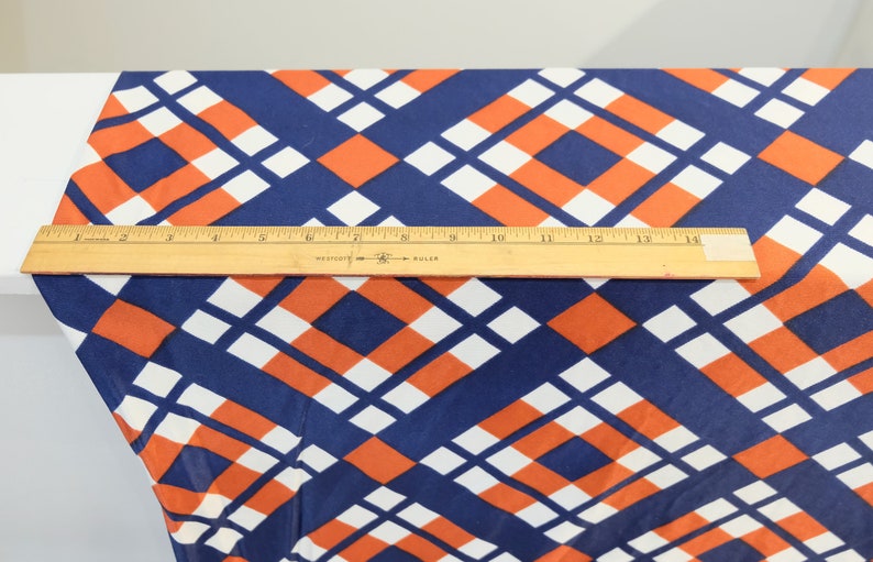 May include: A ruler measuring 14 inches is placed on top of a fabric with a geometric pattern of orange, white, and blue squares.