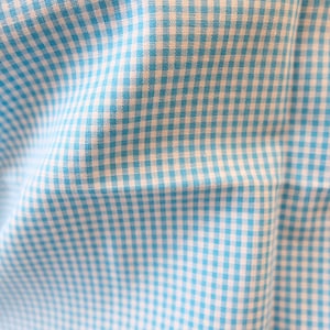 May include: A fabric with a light blue and white gingham check pattern. The material features a small, repeating square grid design with alternating light blue and white colors, showing soft folds and creases across the surface of the textile.
