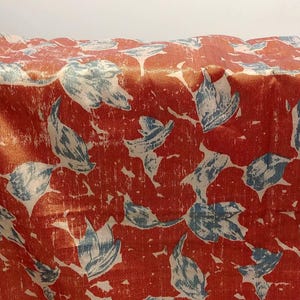 May include: A fabric textile featuring a repeating pattern of stylized blue leaves set against a vibrant burnt orange background. The design displays a distressed, textured appearance with visible folds and creases across the surface of the material.