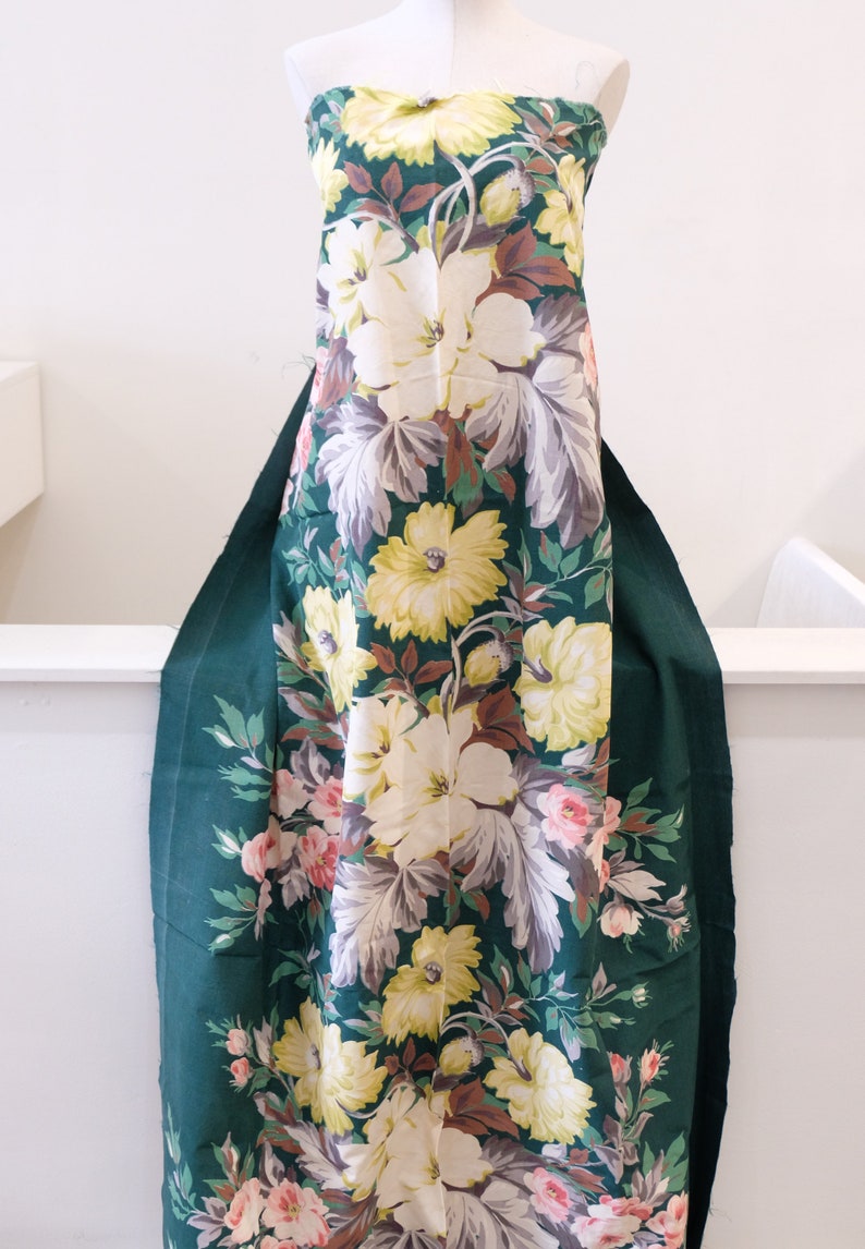 K&ouml;nnte beinhalten: Ein gr&uuml;ner Stoff mit einem gro&szlig;en Blumendruck mit wei&szlig;en, gelben und rosa Bl&uuml;ten. Der Stoff hat einen Vintage-Charakter und w&auml;re perfekt f&uuml;r ein Kleid oder ein anderes Kleidungsst&uuml;ck.