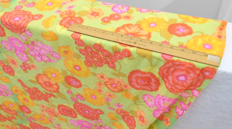 Puede incluir: Una tela verde con estampado floral con grandes flores naranjas, amarillas y rosas. La tela se muestra con una regla para la escala.