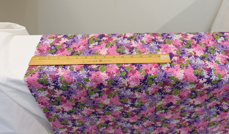 May include: A close-up of a fabric with a floral print. The fabric is a mix of pink, purple, and green. The flowers are small and clustered together. A ruler is placed on top of the fabric to show the scale.