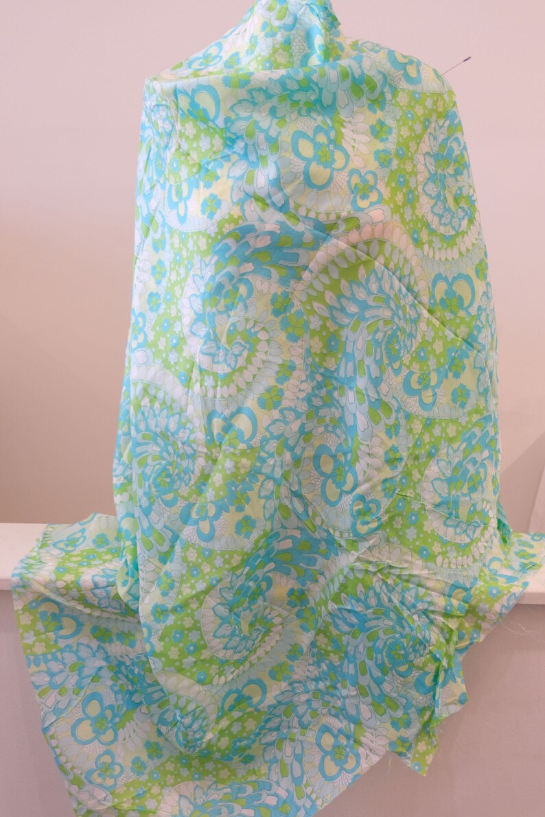 May include: A light blue and green fabric with a swirling floral pattern. The fabric is draped over a form and has a white background.
