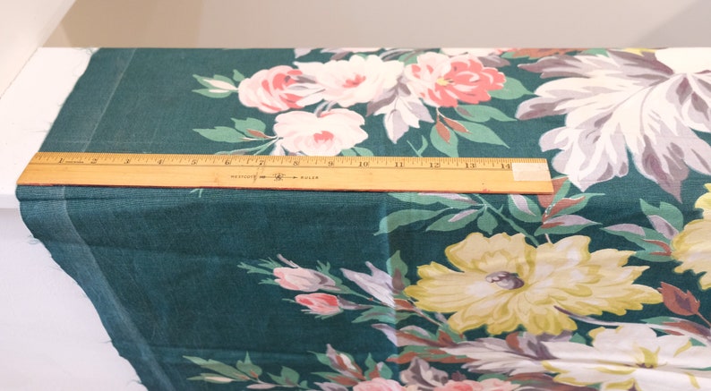 K&ouml;nnte beinhalten: Nahaufnahme eines t&uuml;rkisfarbenen Stoffes mit einem Blumendruck mit gro&szlig;en rosa, wei&szlig;en und gelben Bl&uuml;ten. Ein Holzliniall liegt auf dem Stoff.