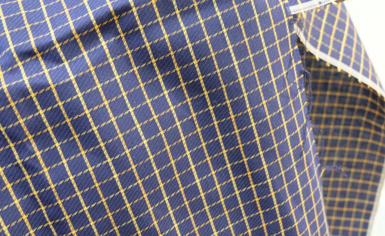 May include: A close-up of a blue and gold plaid fabric. The fabric has a woven texture and a subtle sheen.