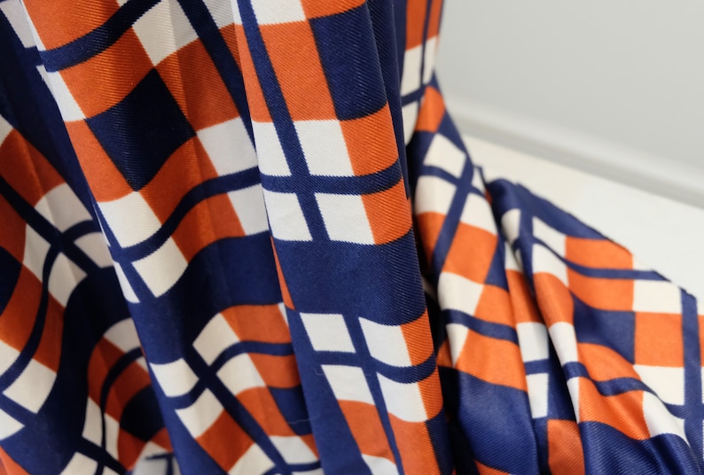 May include: Close-up of a fabric with a geometric pattern in shades of orange, white, and blue. The fabric is folded and the pattern is visible in the folds.