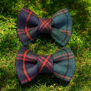May include: Two plaid bow ties in navy blue, teal, and red, resting on green grass. The bow ties have a classic shape and a textured fabric. The plaid pattern features intersecting lines of red against the darker background.