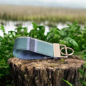 Peut inclure: Un porte-clés tissé avec un motif à carreaux bleu, vert et noir. Le porte-clés est doté d'un fermoir et d'un anneau en métal de couleur dorée. L'objet est posé sur une souche d'arbre, avec un feuillage vert en arrière-plan.