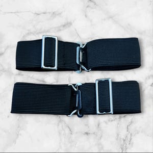 May include: Two black elastic suspenders with silver metal clasps. The suspenders are made of a stretchy fabric and have a simple design.