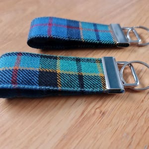 Op de afbeelding: Twee sleutelhangers met zilveren sleutelringen. De sleutelhangers zijn gemaakt van stof met een blauw, groen en geel ruitpatroon.