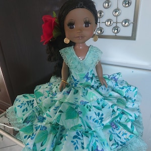 May include: A doll with dark hair and brown eyes, wearing a turquoise dress with a floral and butterfly pattern. The dress has multiple ruffled layers and lace trim. A red flower adorns the doll's hair, and gold earrings are visible.