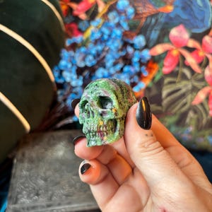 May include: A small, hand-carved skull figurine crafted from a mottled green and deep red mineral stone. The surface features a natural, textured appearance with visible crystalline inclusions throughout the structure of the skull.