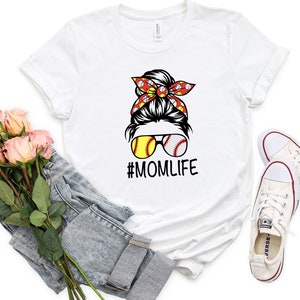 May include: White t-shirt with a graphic of a woman with a messy bun wearing sunglasses with baseballs for lenses and a red bandana with white baseballs. The text "#MOMLIFE" is below the graphic.