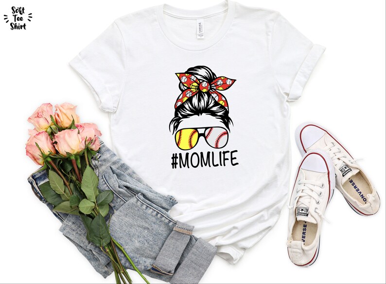 May include: White t-shirt with a graphic of a woman with a messy bun wearing sunglasses with baseballs for lenses and a red bandana with white baseballs. The text "#MOMLIFE" is below the graphic.