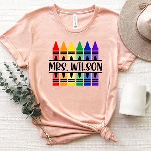 May include: A pink t-shirt with a design of rainbow colored crayons and the text "MRS. WILSON" in black.