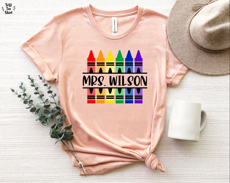 May include: A pink t-shirt with a design of rainbow colored crayons and the text "MRS. WILSON" in black.