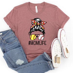 May include: A mauve t-shirt with a graphic of a woman with a bun in her hair wearing sunglasses with baseballs for lenses. The woman is wearing a red and white bandana with baseballs on it. The text "#MOMLIFE" is below the graphic.