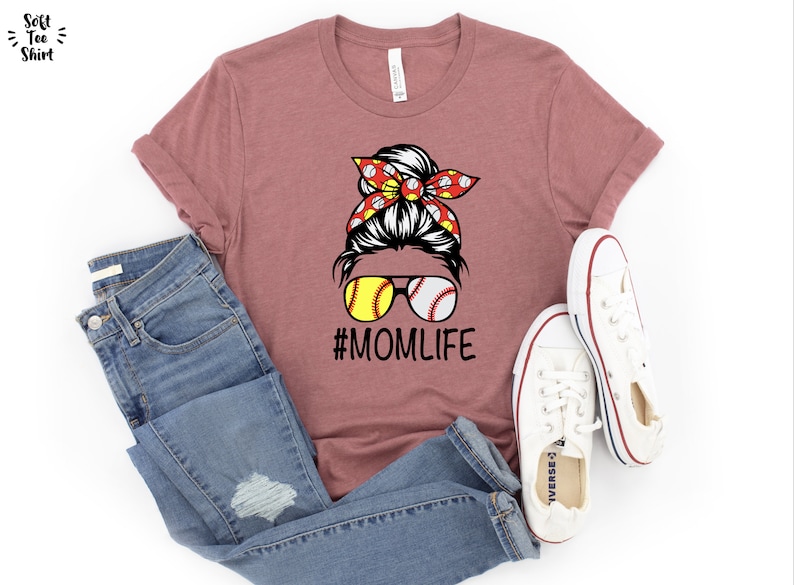 May include: A mauve t-shirt with a graphic of a woman with a bun in her hair wearing sunglasses with baseballs for lenses. The woman is wearing a red and white bandana with baseballs on it. The text "#MOMLIFE" is below the graphic.