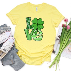 May include: A yellow t-shirt with a green "LOVE" graphic featuring a four-leaf clover. The shirt is displayed on a white background with a pair of white sneakers, blue jeans, a magazine, and a bouquet of pink tulips.