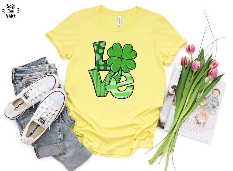 May include: A yellow t-shirt with a green "LOVE" graphic featuring a four-leaf clover. The shirt is displayed on a white background with a pair of white sneakers, blue jeans, a magazine, and a bouquet of pink tulips.