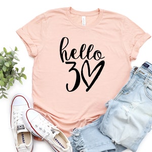 May include: A light pink t-shirt with the words "hello 30" in black script and a heart. The shirt is paired with light blue jeans and white sneakers. A sprig of greenery is also in the image.