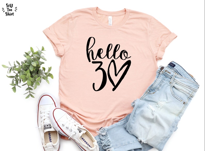 May include: A light pink t-shirt with the words "hello 30" in black script and a heart. The shirt is paired with light blue jeans and white sneakers. A sprig of greenery is also in the image.