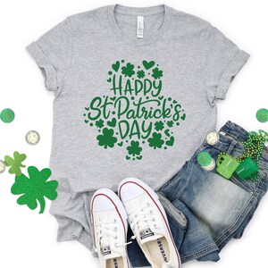 May include: A gray t-shirt with the text "Happy St. Patrick's Day" in green lettering surrounded by shamrocks and hearts. The shirt is laid out on a white surface with a pair of white sneakers, blue jeans, and green shamrock decorations.