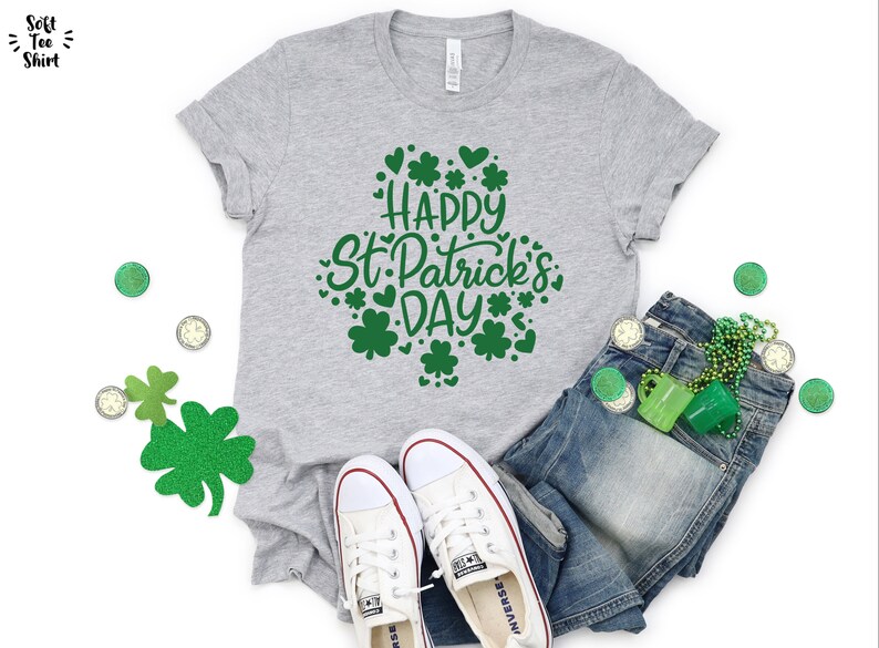 May include: A gray t-shirt with the text "Happy St. Patrick's Day" in green lettering surrounded by shamrocks and hearts. The shirt is laid out on a white surface with a pair of white sneakers, blue jeans, and green shamrock decorations.