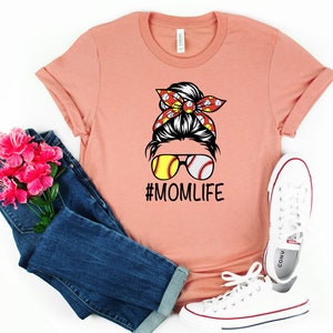May include: A peach colored t-shirt with a graphic of a woman with a messy bun, wearing sunglasses with baseballs for lenses. The bun is decorated with a red and white bandana with baseballs. The text "#MOMLIFE" is printed below the graphic.