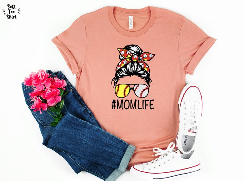 May include: A peach colored t-shirt with a graphic of a woman with a messy bun, wearing sunglasses with baseballs for lenses. The bun is decorated with a red and white bandana with baseballs. The text "#MOMLIFE" is printed below the graphic.