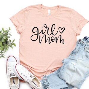 May include: A light pink t-shirt with the text "girl mom" in black script with a small heart symbol.