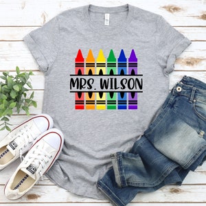 May include: Gray t-shirt with a rainbow of crayons and the text "MRS. WILSON" printed on it. The shirt is displayed on a wooden surface with a pair of white sneakers and blue jeans.