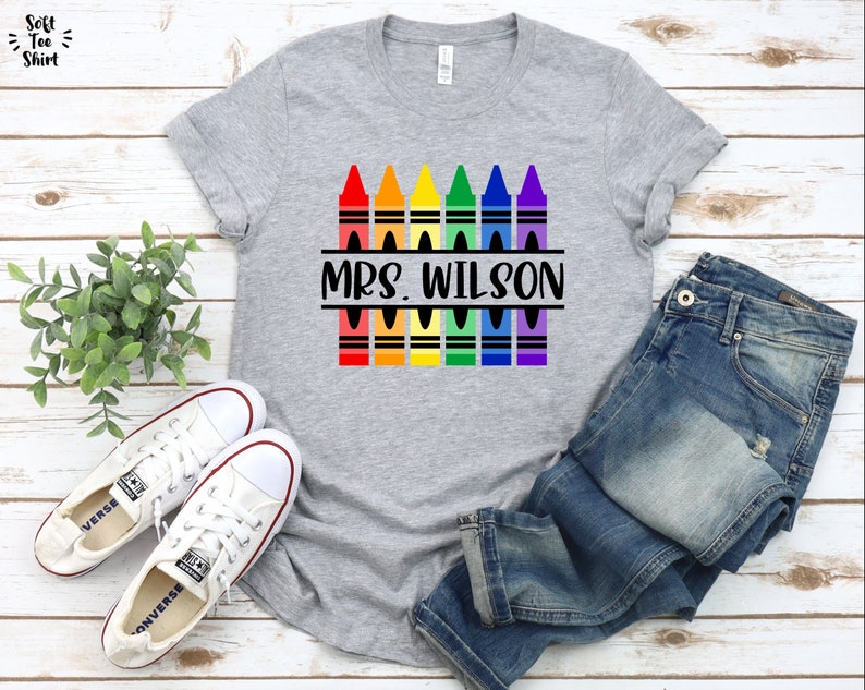 May include: Gray t-shirt with a rainbow of crayons and the text "MRS. WILSON" printed on it. The shirt is displayed on a wooden surface with a pair of white sneakers and blue jeans.