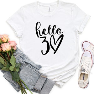 May include: White t-shirt with the text "hello 30" in black script and a heart. The shirt is paired with a pair of light-wash denim jeans, a bouquet of pink roses, and white sneakers.