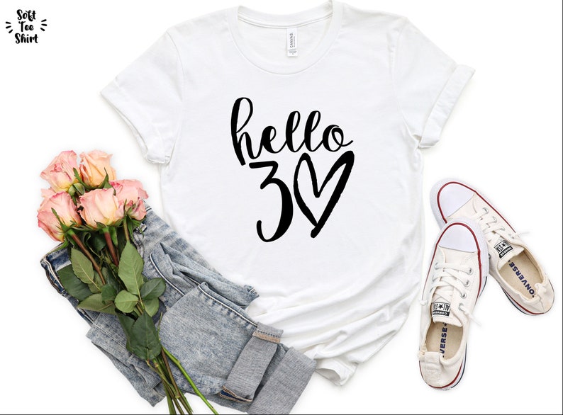 May include: White t-shirt with the text "hello 30" in black script and a heart. The shirt is paired with a pair of light-wash denim jeans, a bouquet of pink roses, and white sneakers.