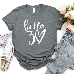 May include: A heather gray t-shirt with the text "hello 30" in white script and a heart. The shirt is paired with white sneakers and light-wash denim jeans. A bouquet of pink flowers adds a decorative touch.