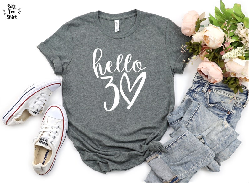 May include: A heather gray t-shirt with the text "hello 30" in white script and a heart. The shirt is paired with white sneakers and light-wash denim jeans. A bouquet of pink flowers adds a decorative touch.