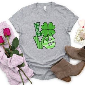 May include: A gray t-shirt with a green "LOVE" graphic featuring a four-leaf clover. The shirt is displayed on a white background with a pair of brown boots, a pink sweater, and a small gift bag.